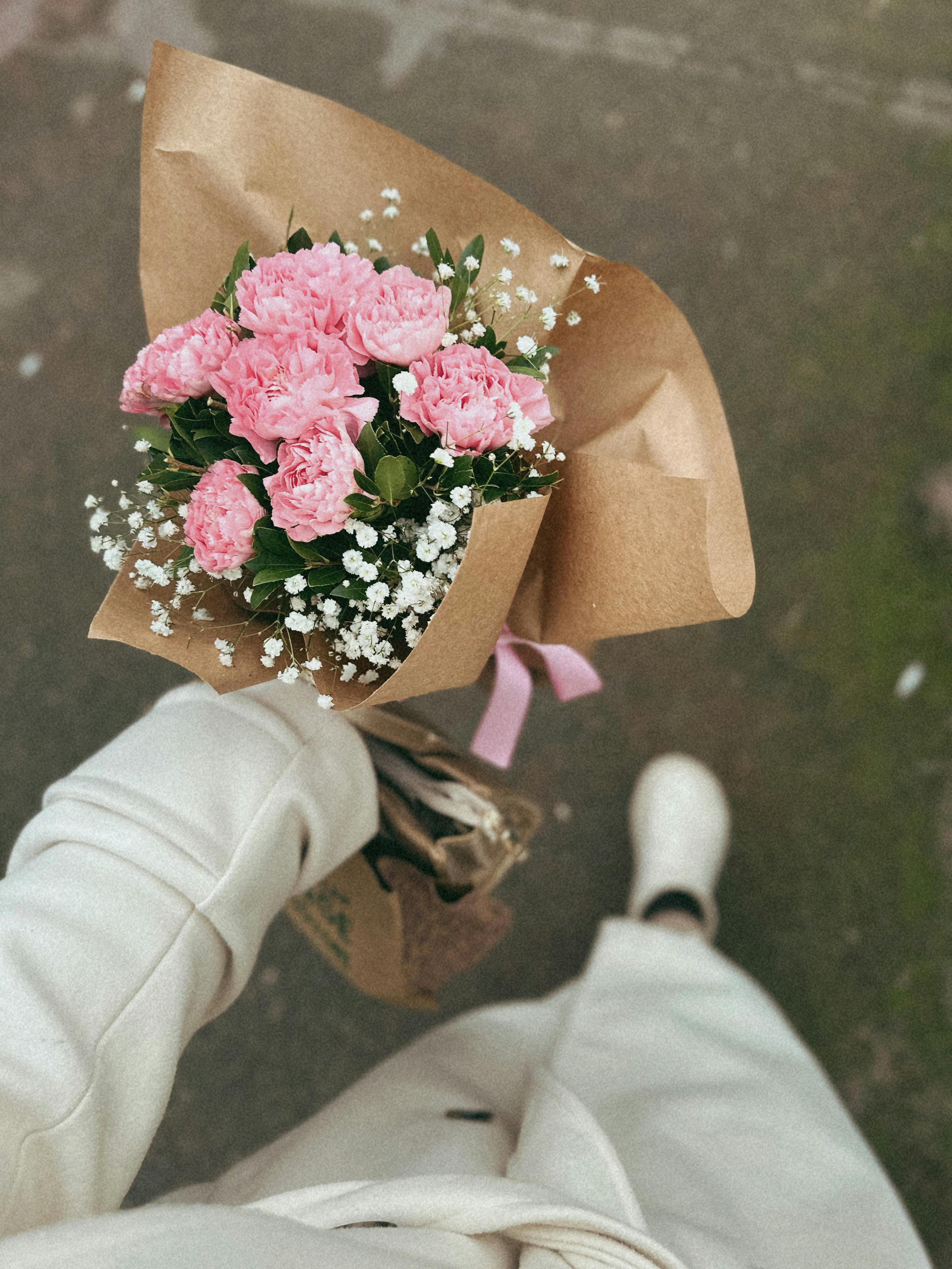 A fashionably dressed woman holds a bouquet of pink carnations wrapped in brown paper.
