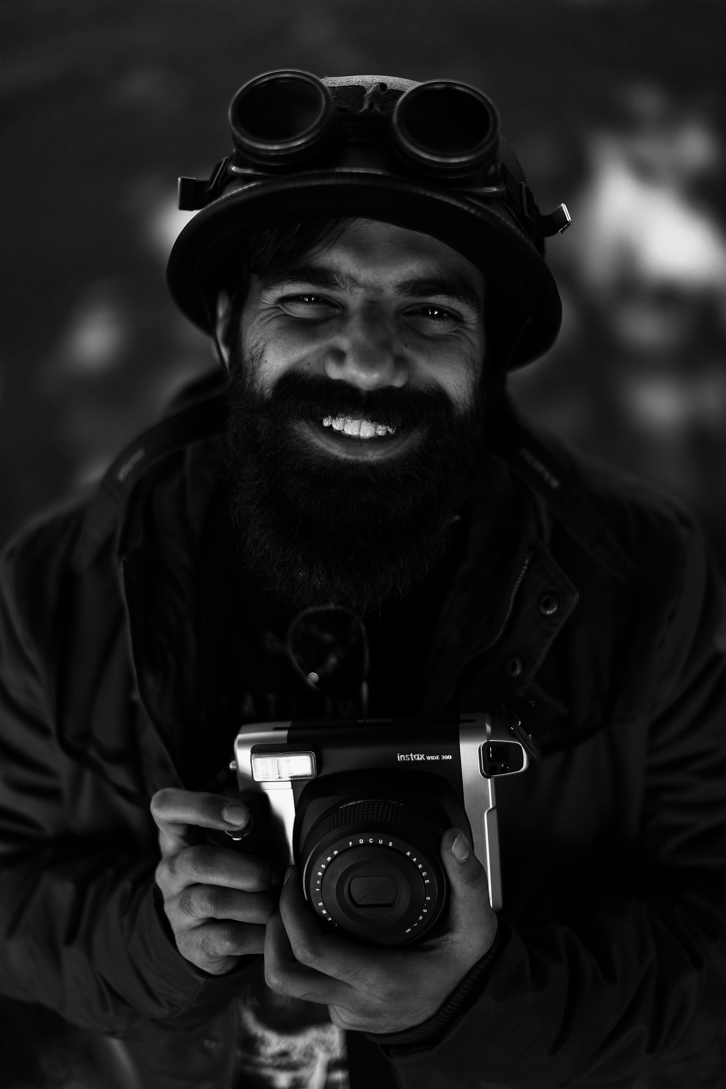 这是一幅单色肖像，画中一位留着胡子的男人戴着老式护目镜，手持相机，面带微笑