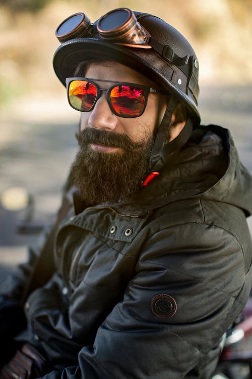 Close-up Photo of Bearded Man in Black Motorcycle Helmet with Brass Goggles, Sunglasses, and Black Hooded Jacket