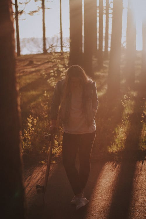 Woman in White Shirt, Blue Denim Jacket and Black Leggings Standing Under Trees Leaning on Skateboard in Hand