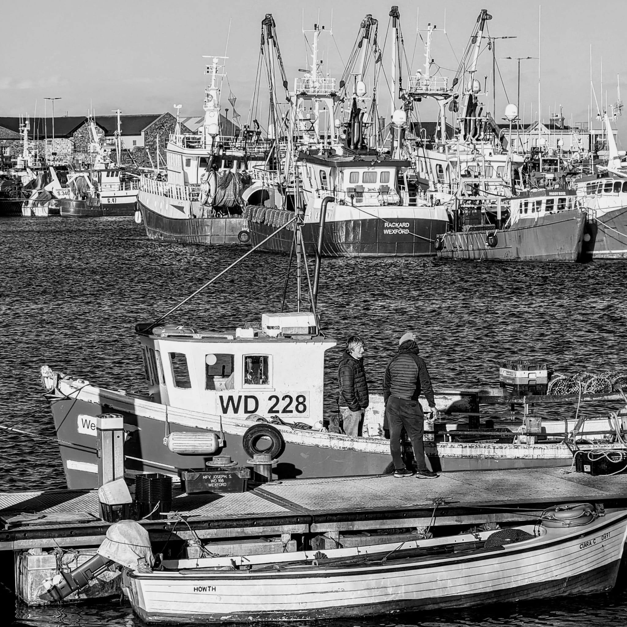 Fishermen prepare their boat at a busy harbor, surrounded by larger vessels.