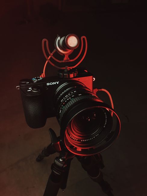 A Sony camera with lens and microphone mounted on a tripod, illuminated by red lighting.