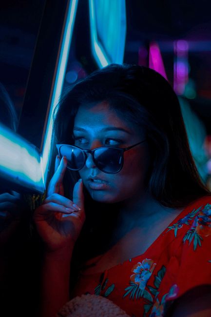 A contemplative woman with sunglasses stands beside a neon light, creating a moody atmosphere.
