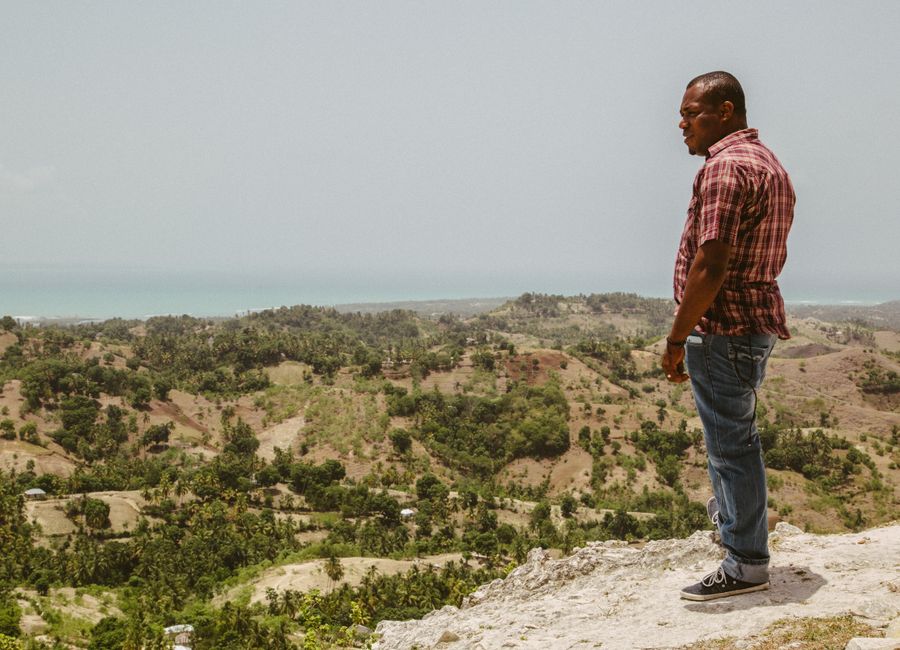 A man stands on a cliff in Haiti, overlooking a scenic landscape with the ocean in the background.