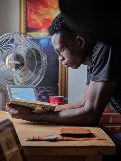 A man reads attentively at a desk in a cozy indoor setting with books and a laptop in Ibadan, Nigeria.