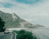 一辆汽车沿着沿海公路行驶，一边是郁郁葱葱的山脉，一边是海景，天空明亮