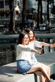 Two Asian women enjoying a sunny day, taking a selfie together by a pond in the city.