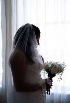 Silhouette of a bride in veil holding a bouquet by a window, exuding elegance and romantic charm.