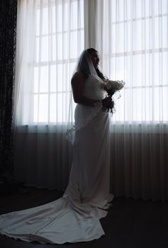 Bride holding bouquet in silhouette by window in wedding dress and veil.
