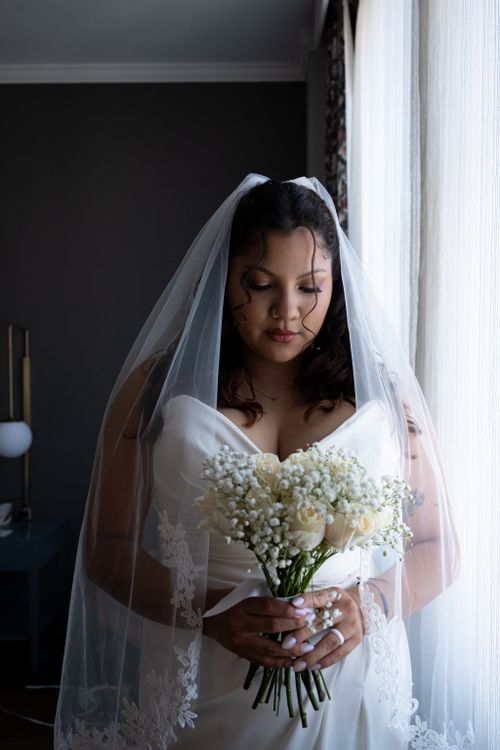 Free Beautiful bride in white dress holding a bouquet, standing by the window with soft lighting. Stock Photo