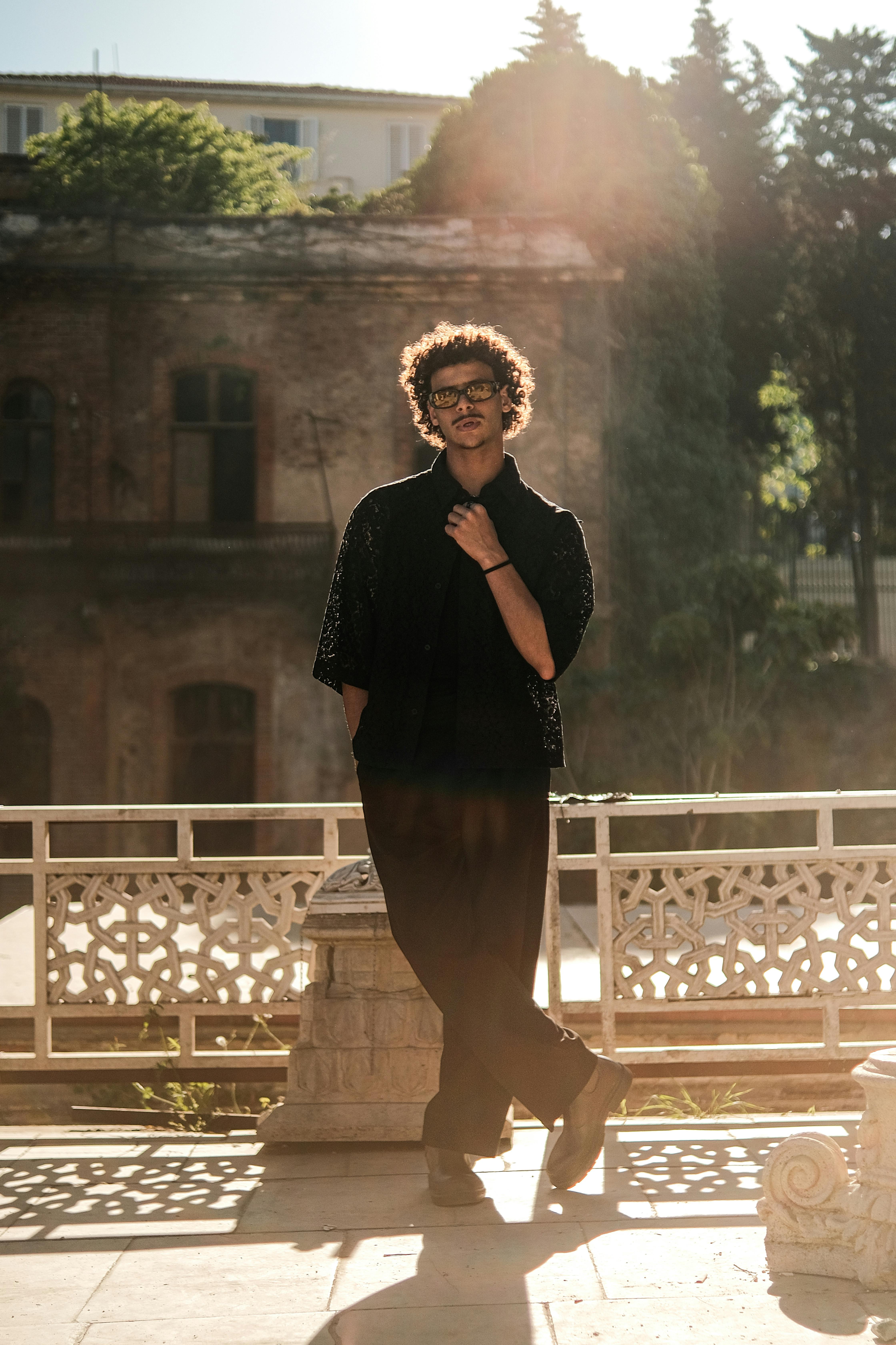 A fashionable man standing outdoors in an urban area with sunbeams highlighting his silhouette, creating a moody scene.