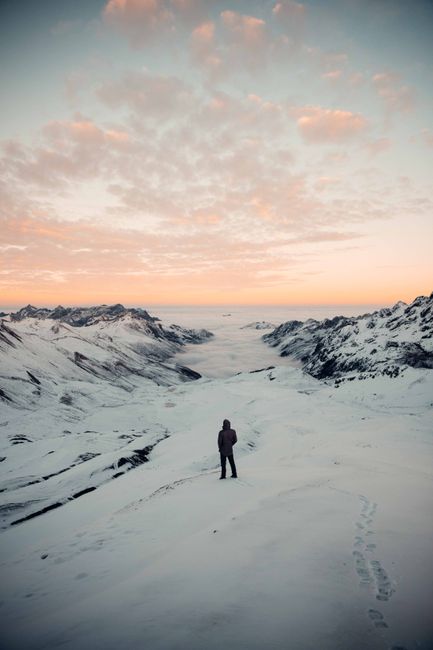 A lone hiker stands on a snow-covered mountain ridge at sunrise, surrounded by a breathtaking winter landscape.