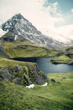 A hiker stands on a cliff overlooking a serene mountain lake surrounded by lush greenery and snow-capped peaks.
