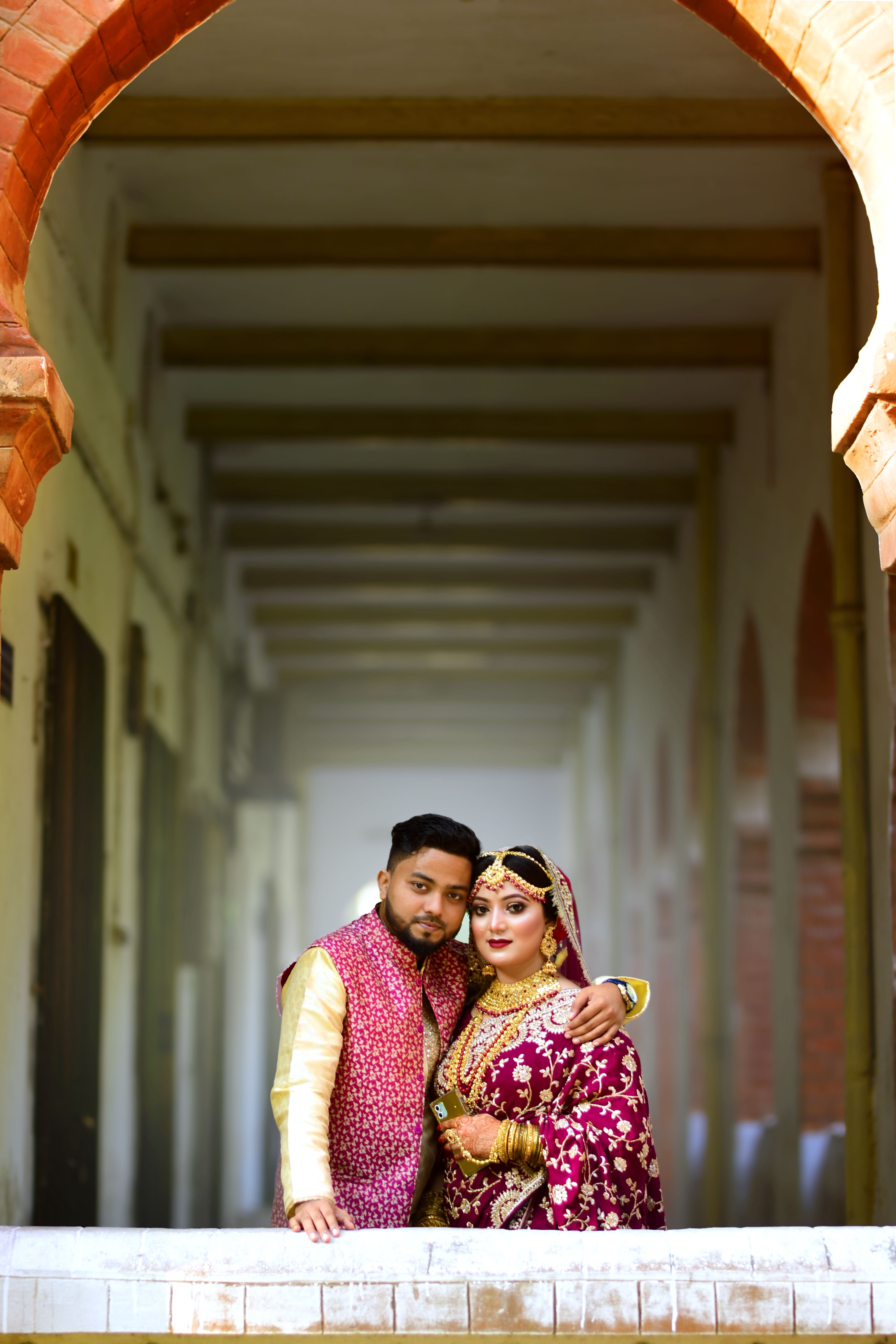 Indian couple in traditional wedding outfits posing under a historic archway.
