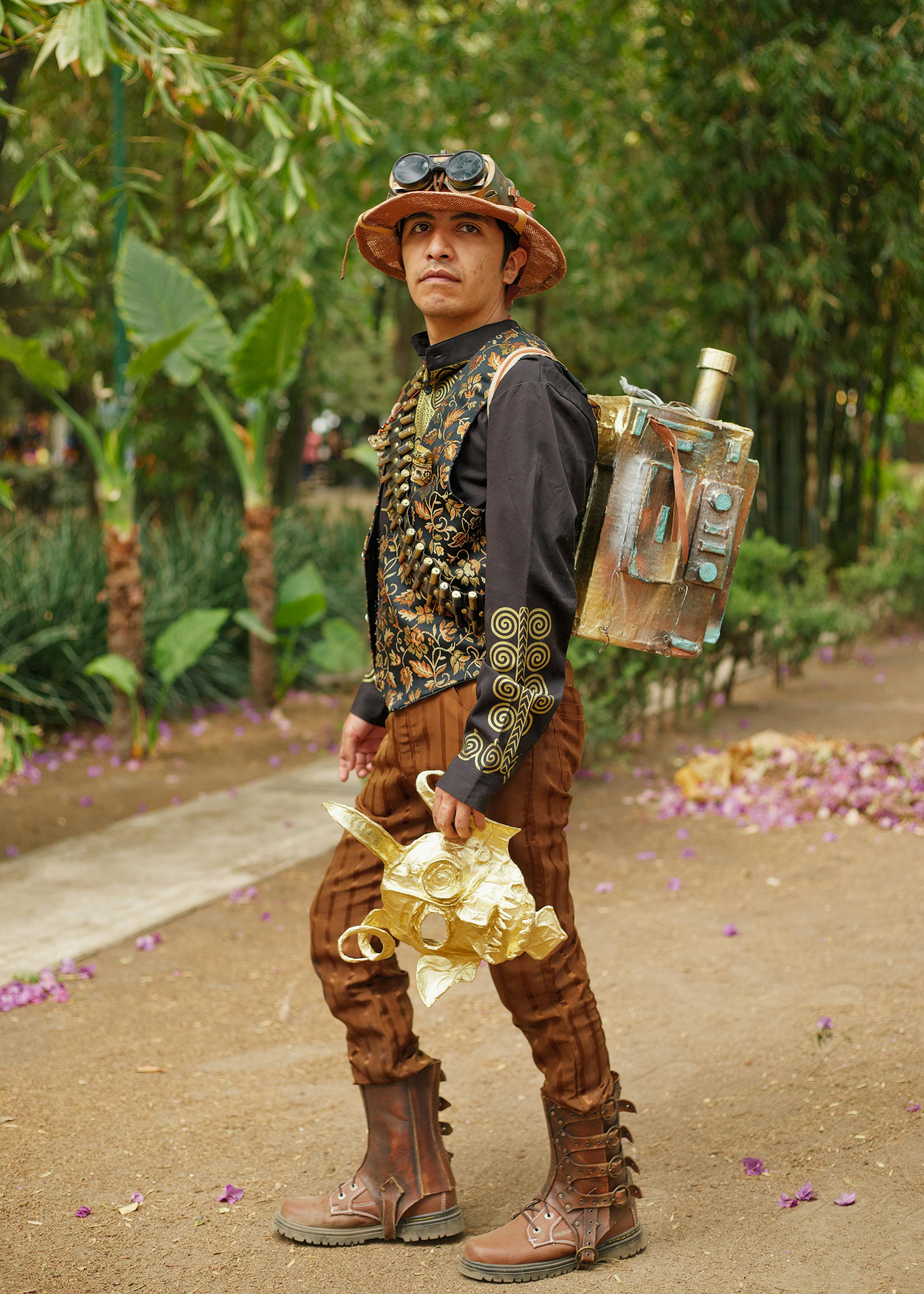A man in steampunk attire poses on a path in a lush tropical garden.
