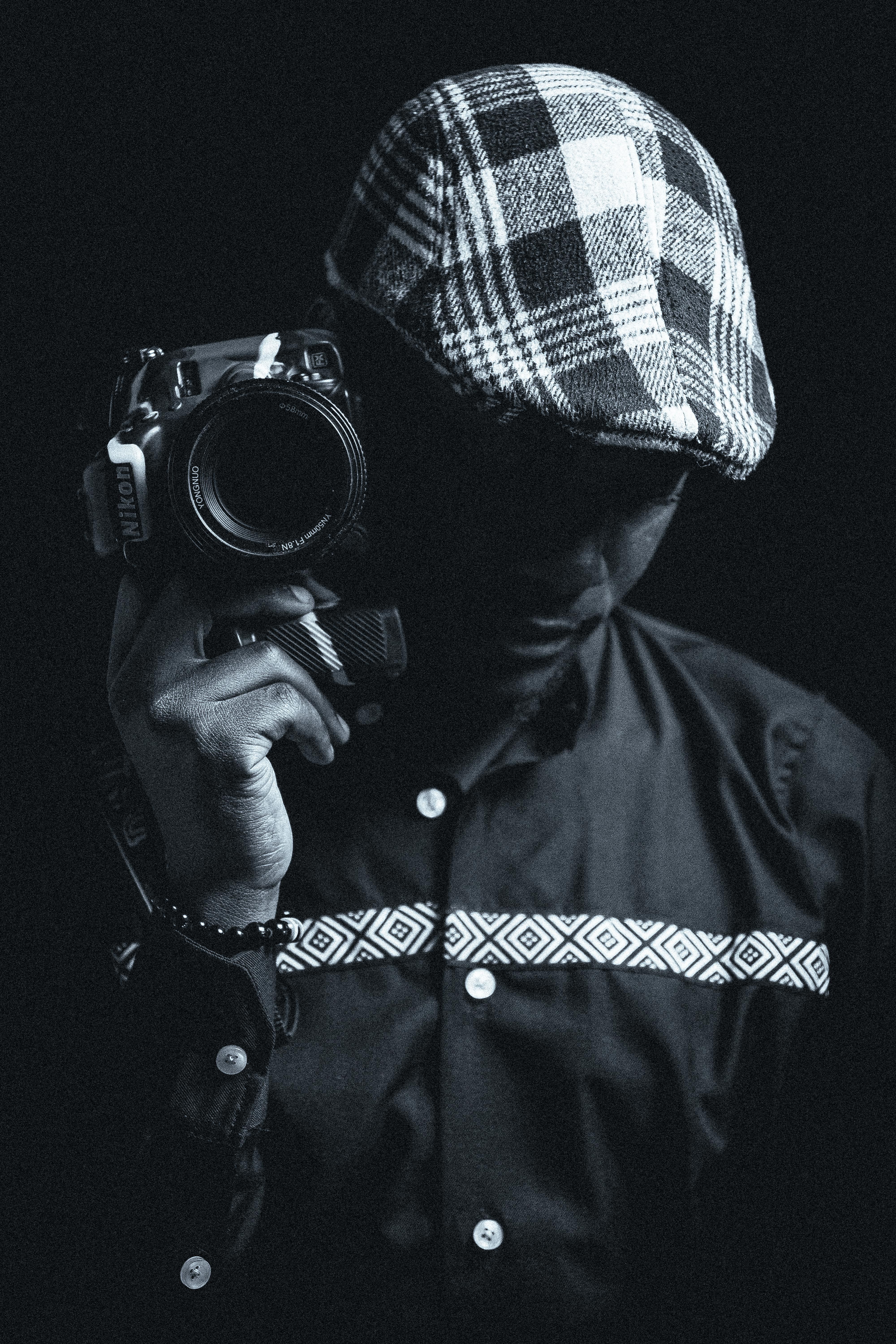 Artistic black and white portrait of a man holding a camera, wearing a plaid beret and patterned shirt in a studio.