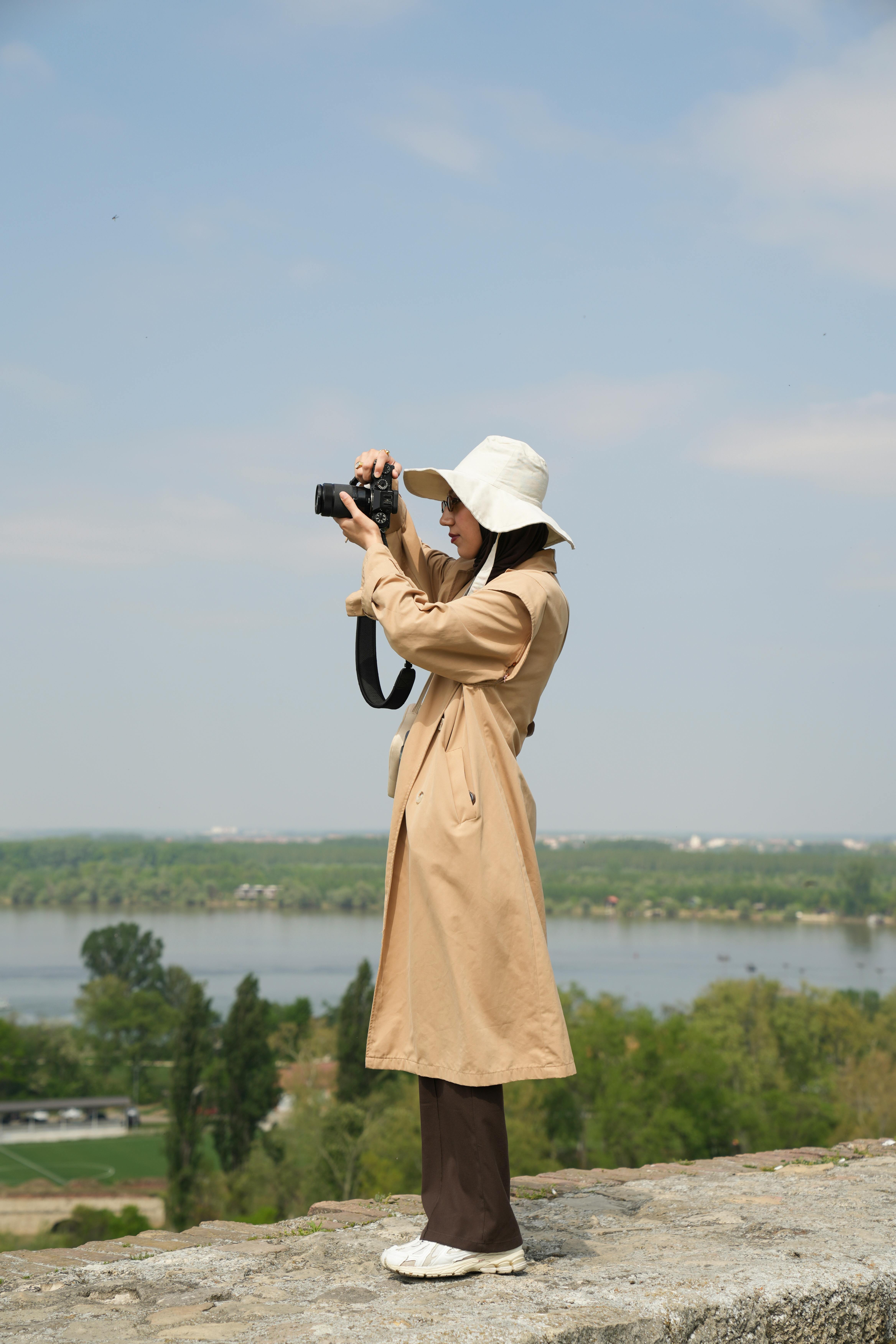 A woman in a coat and hat stands outdoors taking photos with a camera.