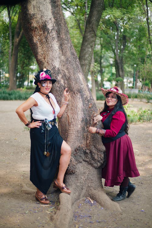 Two women in costumes posing near a tree