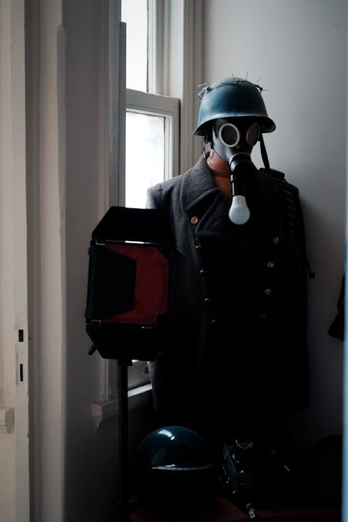 A mannequin wearing a helmet and gas mask stands in front of a window
