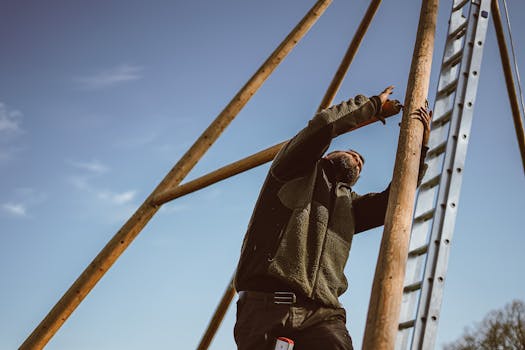 Man climbing ladder and assembling wooden structure against a clear blue sky.