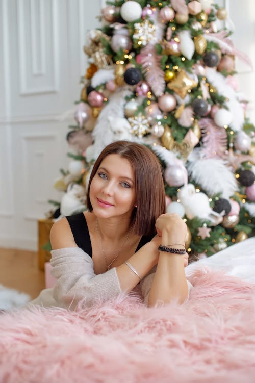 A woman sitting on a pink fur rug in front of a christmas tree