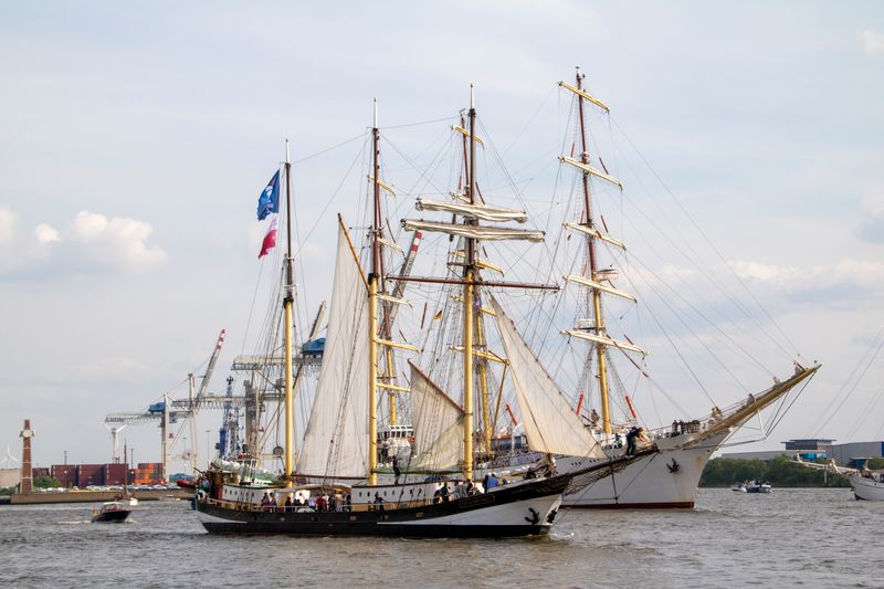 Tall ships with large full sails sailing near a port on a river at the heart of the parade route