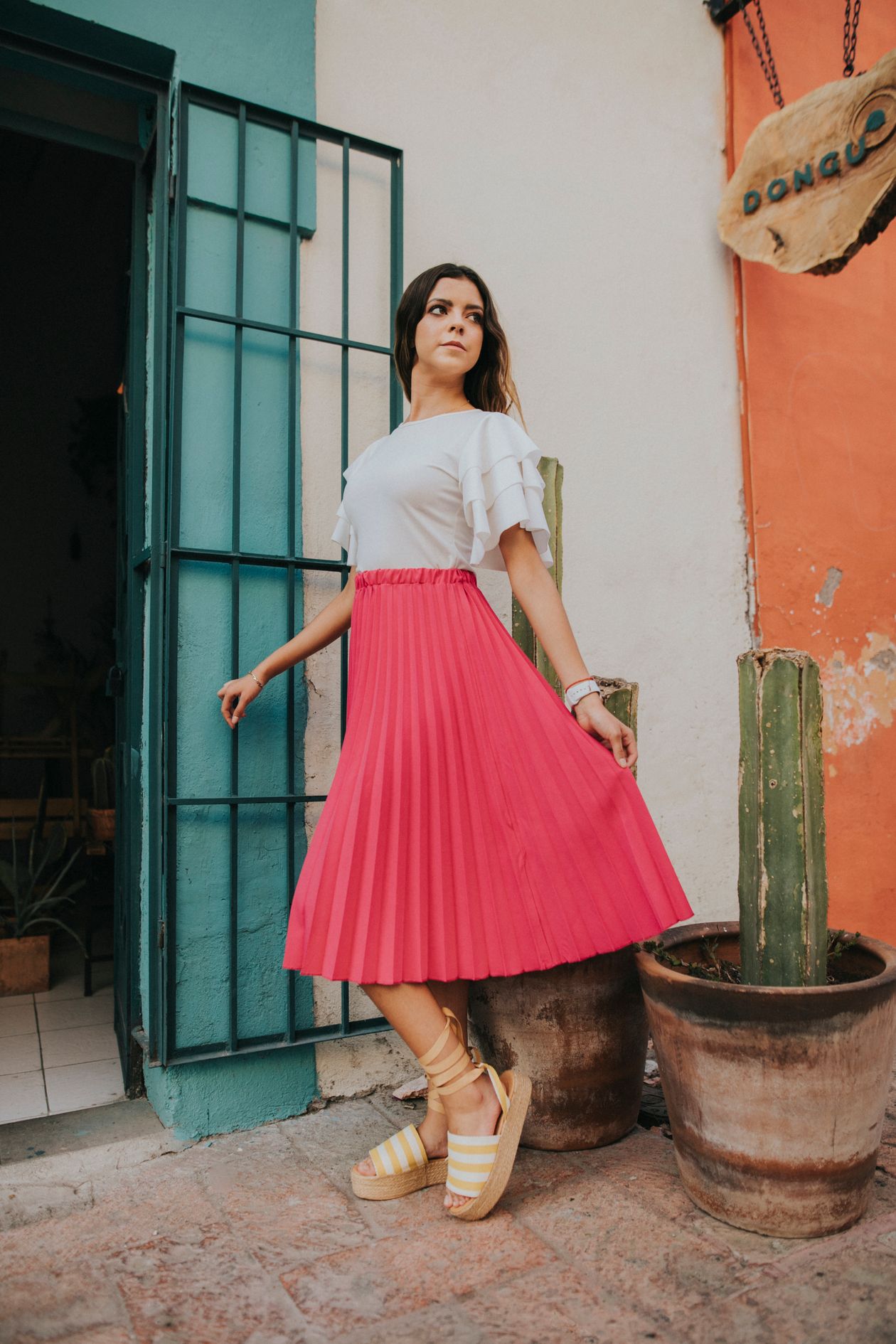 Woman in a white ruffled top and pink pleated midi skirt with striped espadrille sandals, by a doorway with a wooden sign.