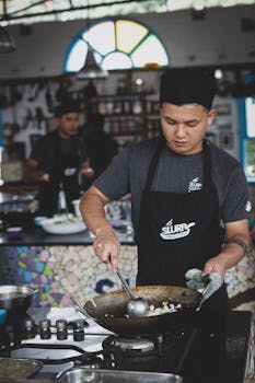 An Asian chef diligently cooking in a restaurant kitchen, wearing a hat and apron.