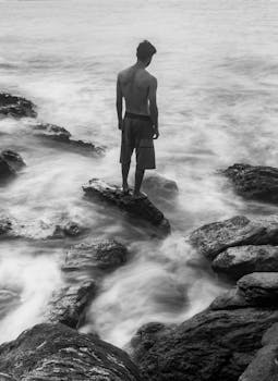 Shirtless man stands on rocky shore, looking out to sea in a black and white photo.