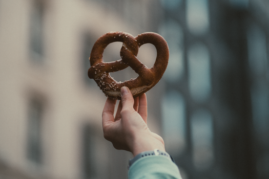 A hand holds a salted pretzel against a blurred urban background, showcasing street food.