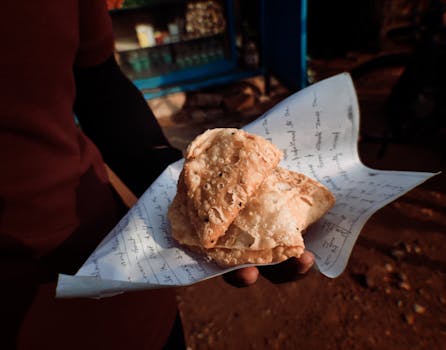 A close-up of a person holding freshly baked samosas wrapped in printed paper, captured outdoors.