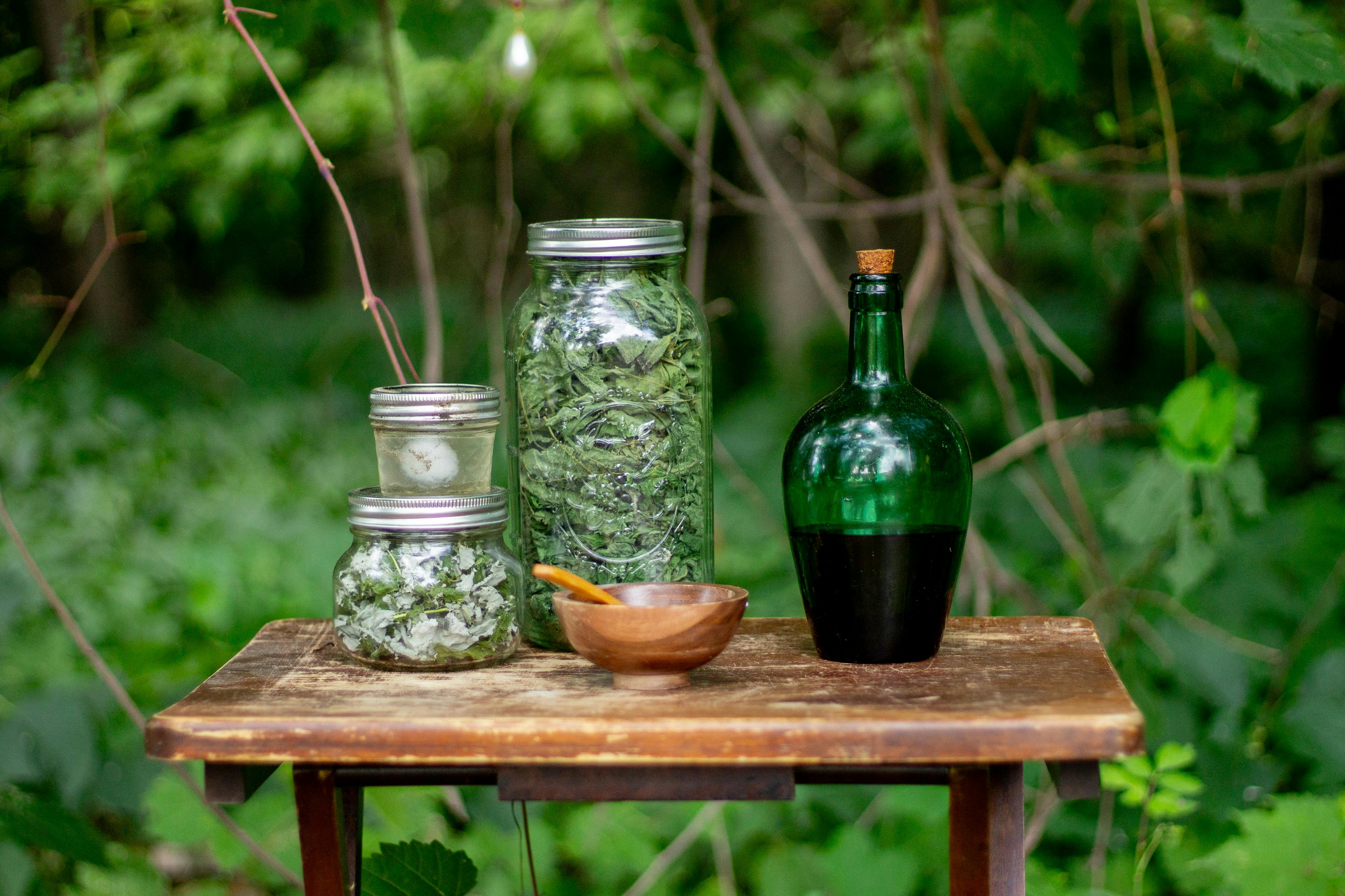 Glass jars of dried herbs and a green bottle on a wooden table outdoors.