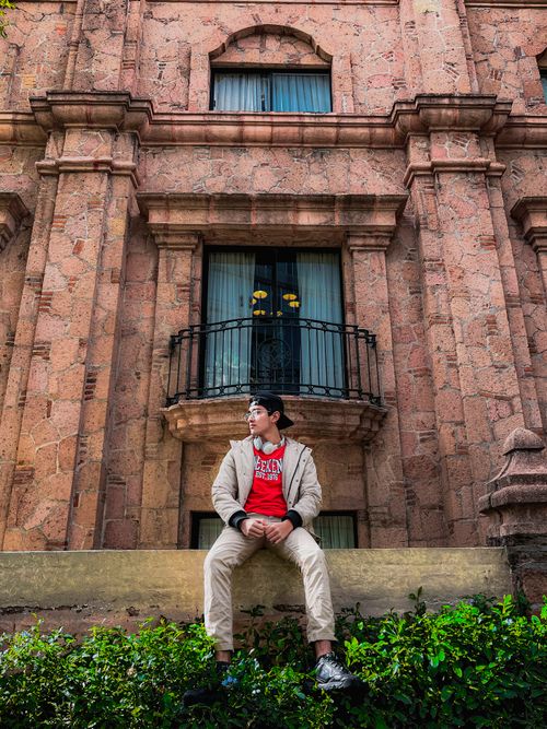 Free Teen wearing a jacket and eyeglasses sitting by an historic building. Stock Photo