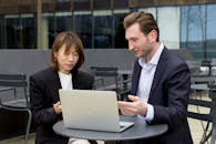 Smiling Businessman and Businesswoman Looking at a Laptop and Talking at a Table in Front of an Office Building