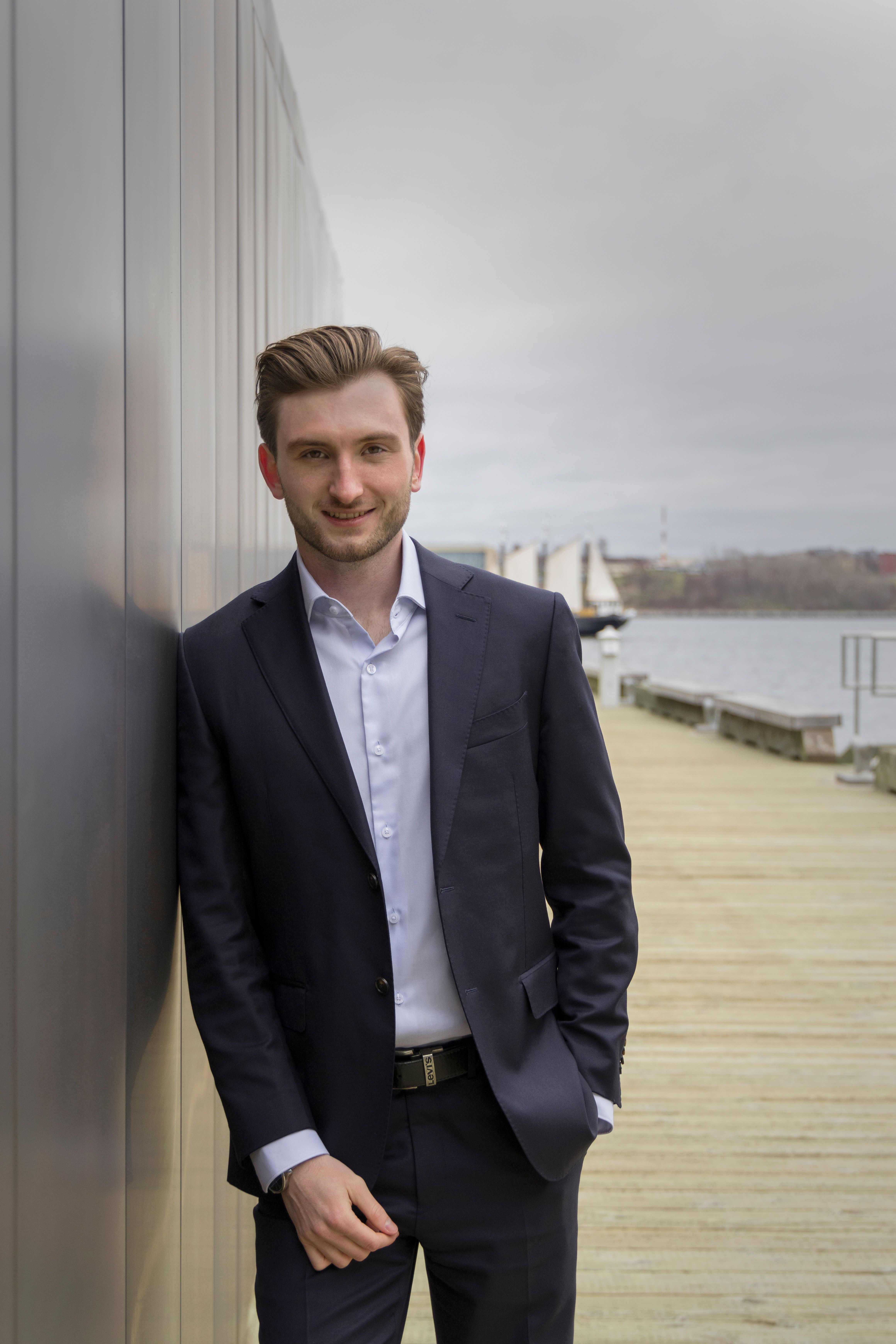 Young businessman in a suit leaning against wall by a lakeside pier, exuding confidence.