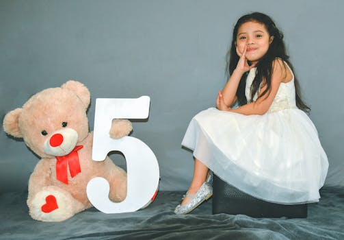 Charming portrait of a girl in a white dress celebrating her fifth birthday with a teddy bear. by Noé Villalta Photography