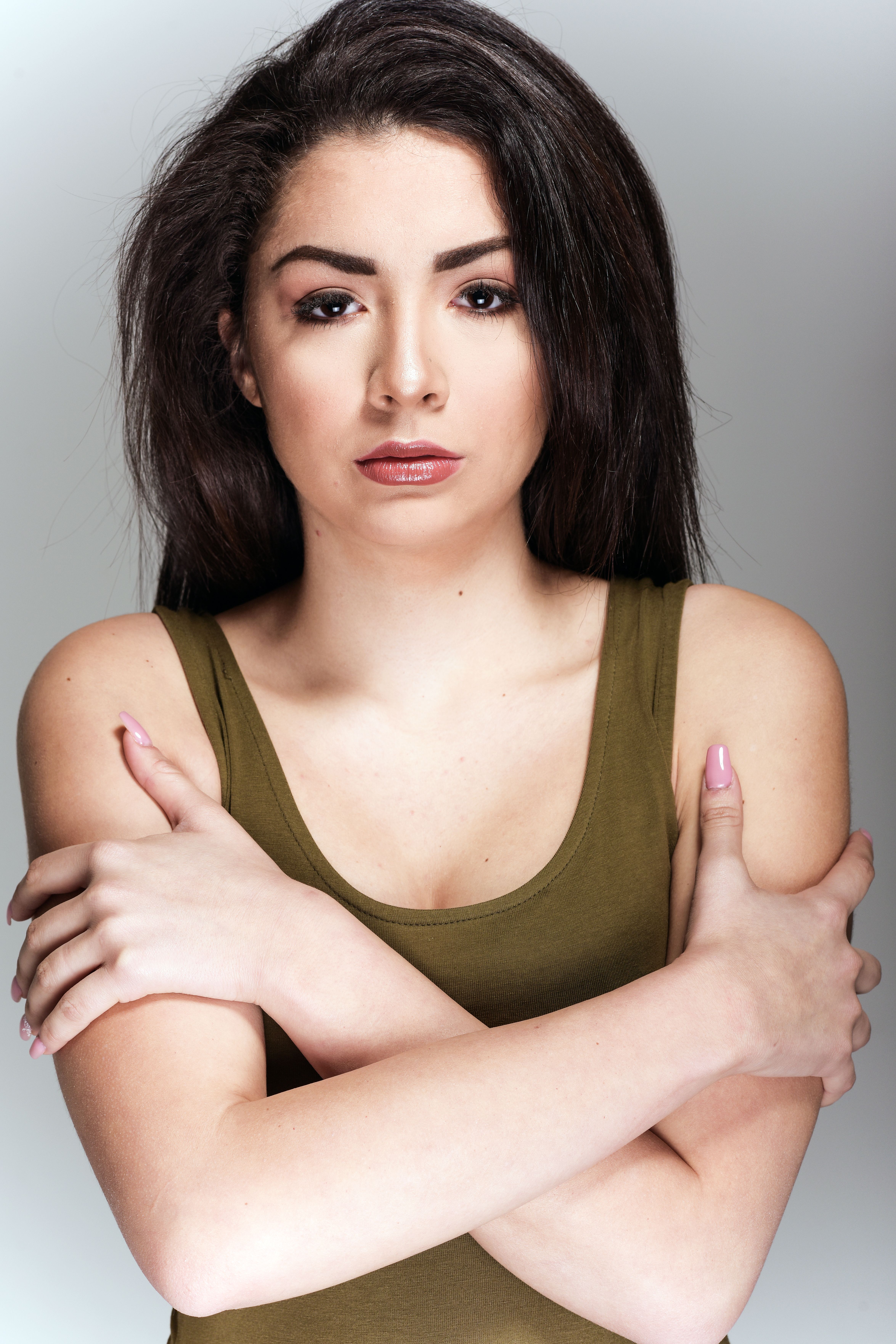 Striking portrait of a brunette woman with makeup, confidently posing in a studio setting, arms crossed.