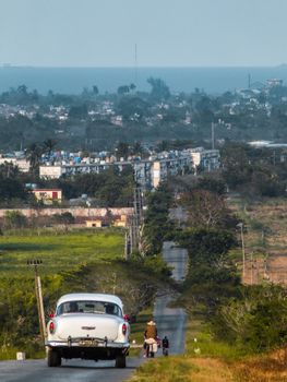 A vintage car travels a rural road in Cárdenas, Cuba, with lush greenery and distant cityscape.