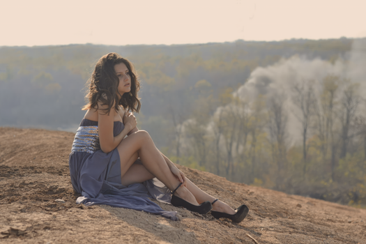 A woman in a glamorous dress sits elegantly on a hilltop amid fall foliage, bathed in sunlight.