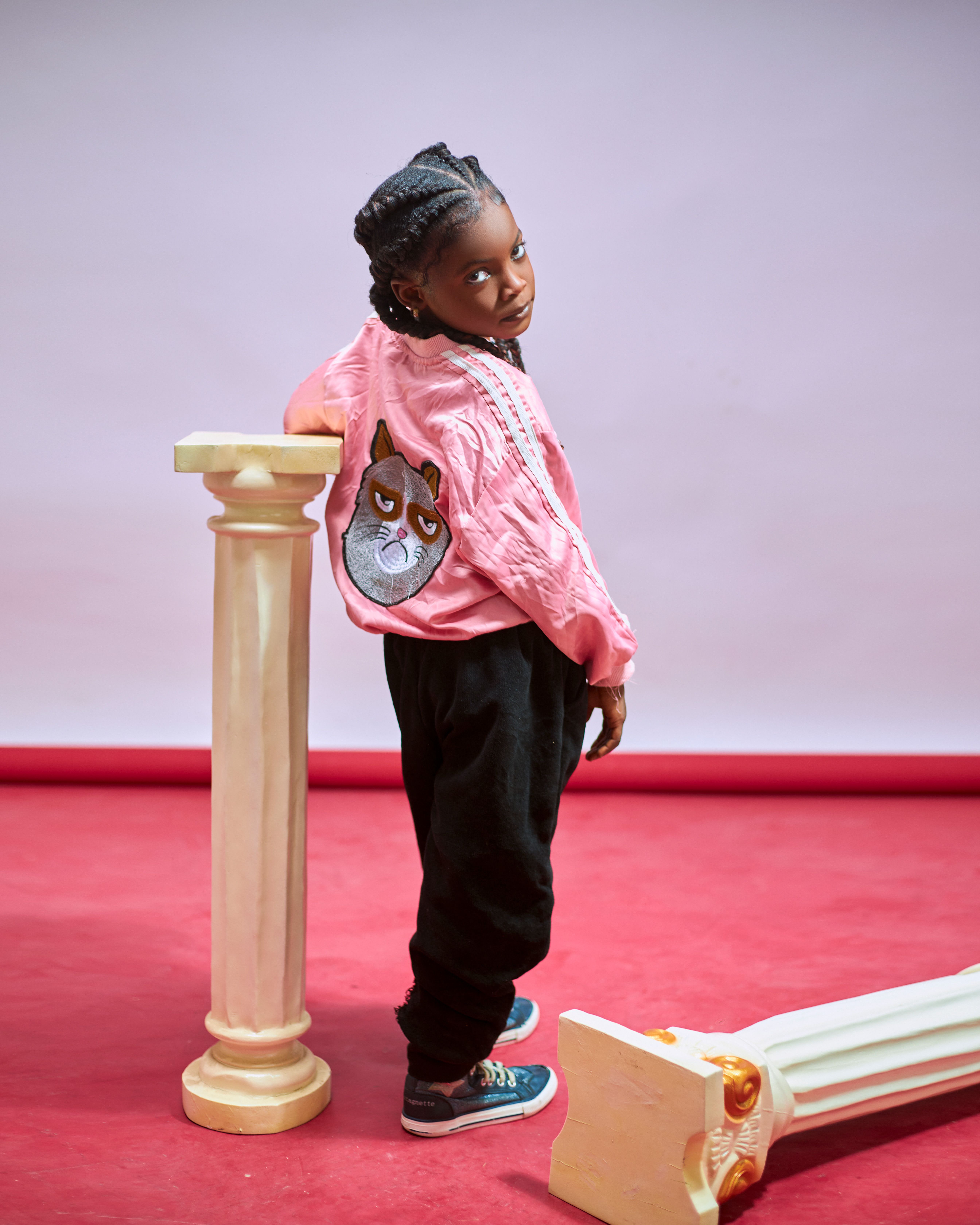 A fashionable child in pink jacket posing confidently next to decorative columns in a studio setting.