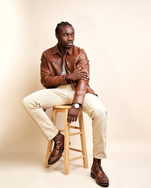 A man in a brown leather jacket and tan pants sitting on a stool