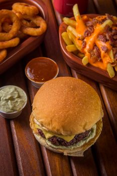 Tasty burger with toppings, cheesy fries, and onion rings on a wooden table, perfect for comfort food lovers.