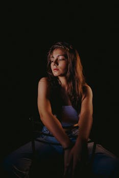 A dramatic portrait of a woman sitting on a chair in a dark studio setting.
