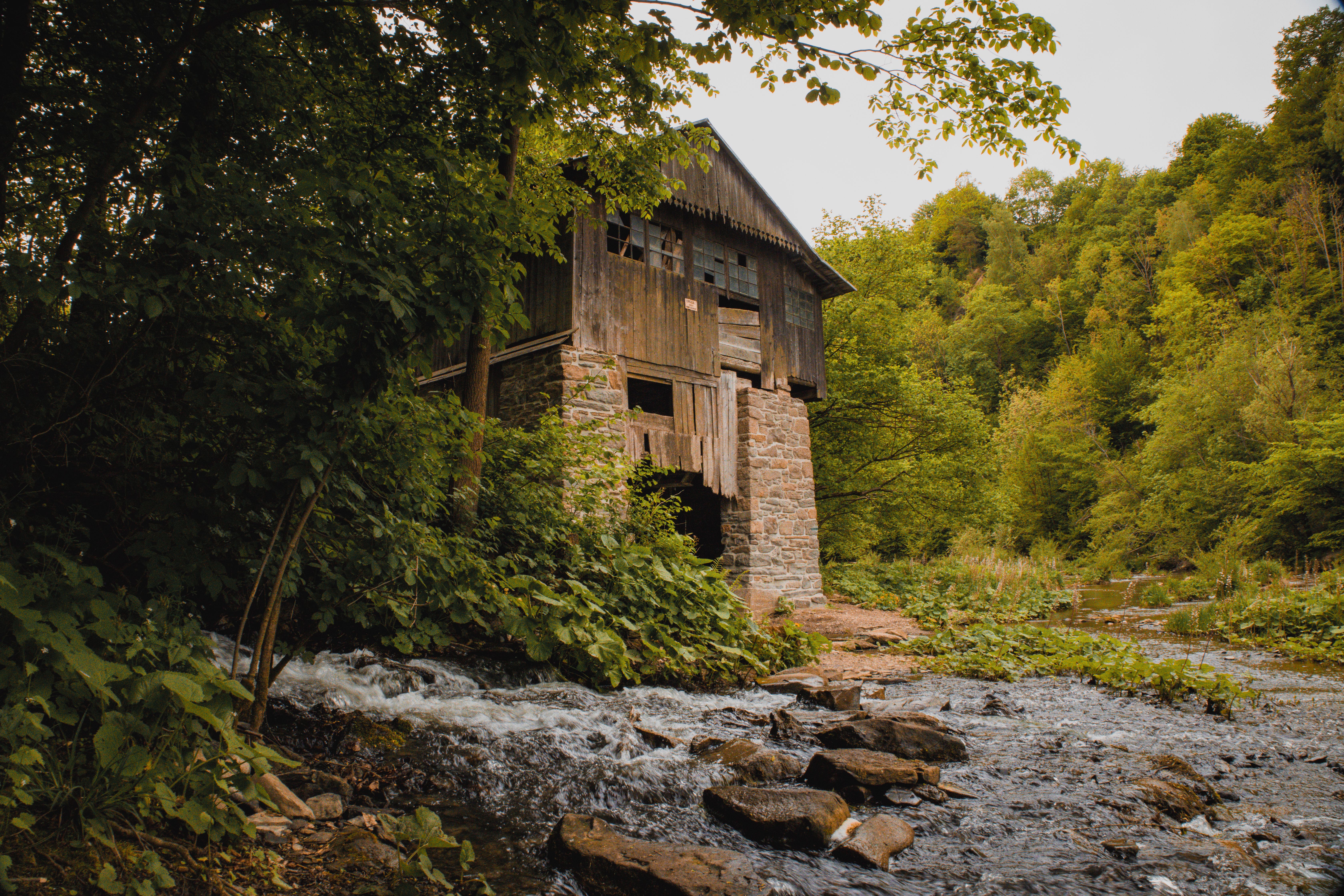 A rustic abandoned wooden mill surrounded by lush forest and a flowing stream under a cloudy sky.