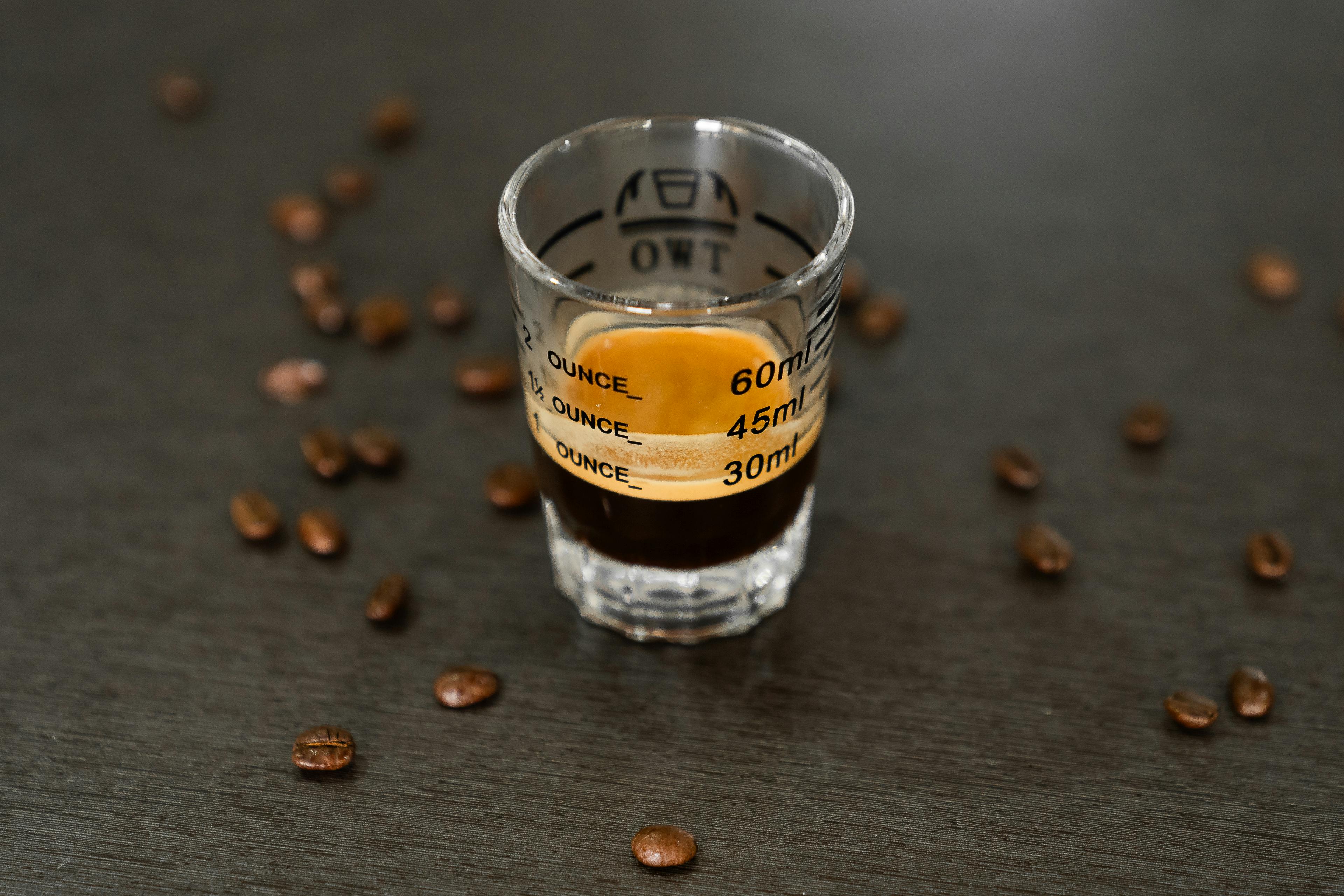 A rich espresso shot in a measuring glass surrounded by scattered coffee beans, perfect for coffee lovers.