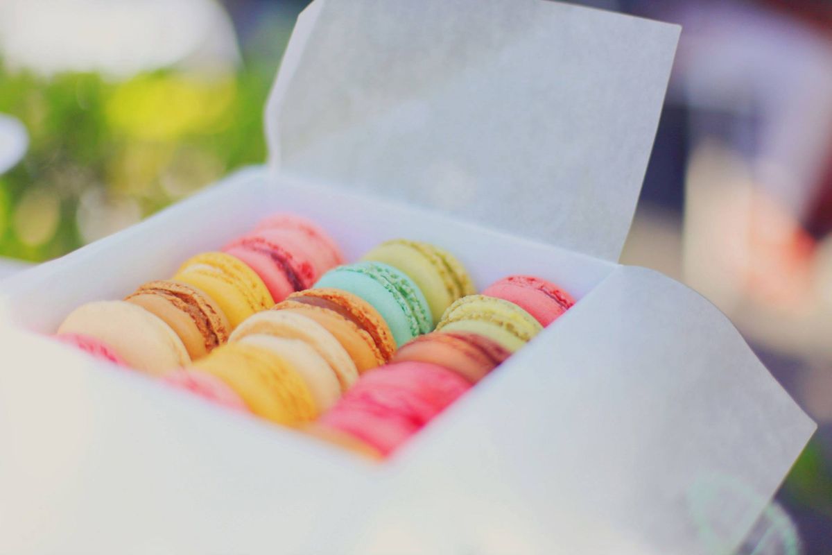 An open white takeaway box of small, colorful macarons in soft natural light.