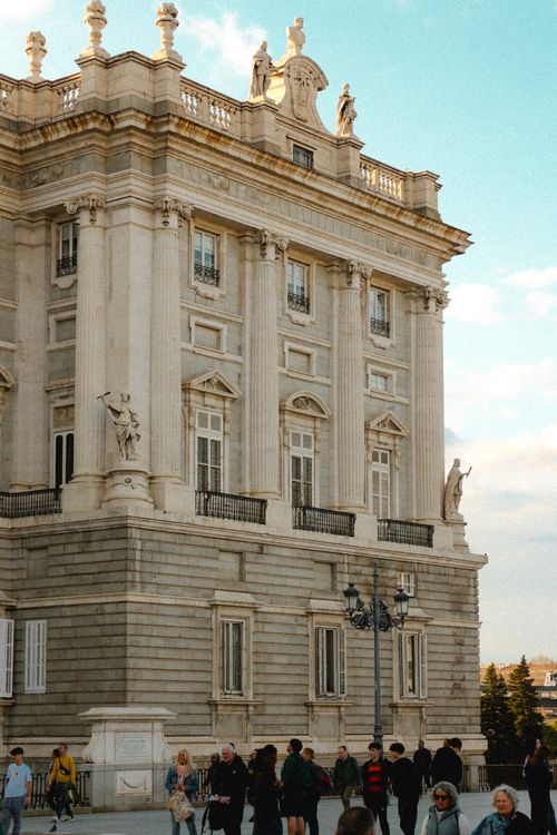 Free View of the Royal Palace of Madrid with tourists exploring the historic architecture in the afternoon light. Stock Photo