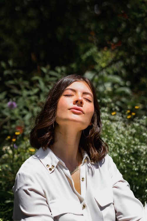 Free Portrait of a woman in a white shirt enjoying sunlight in a lush garden. Stock Photo