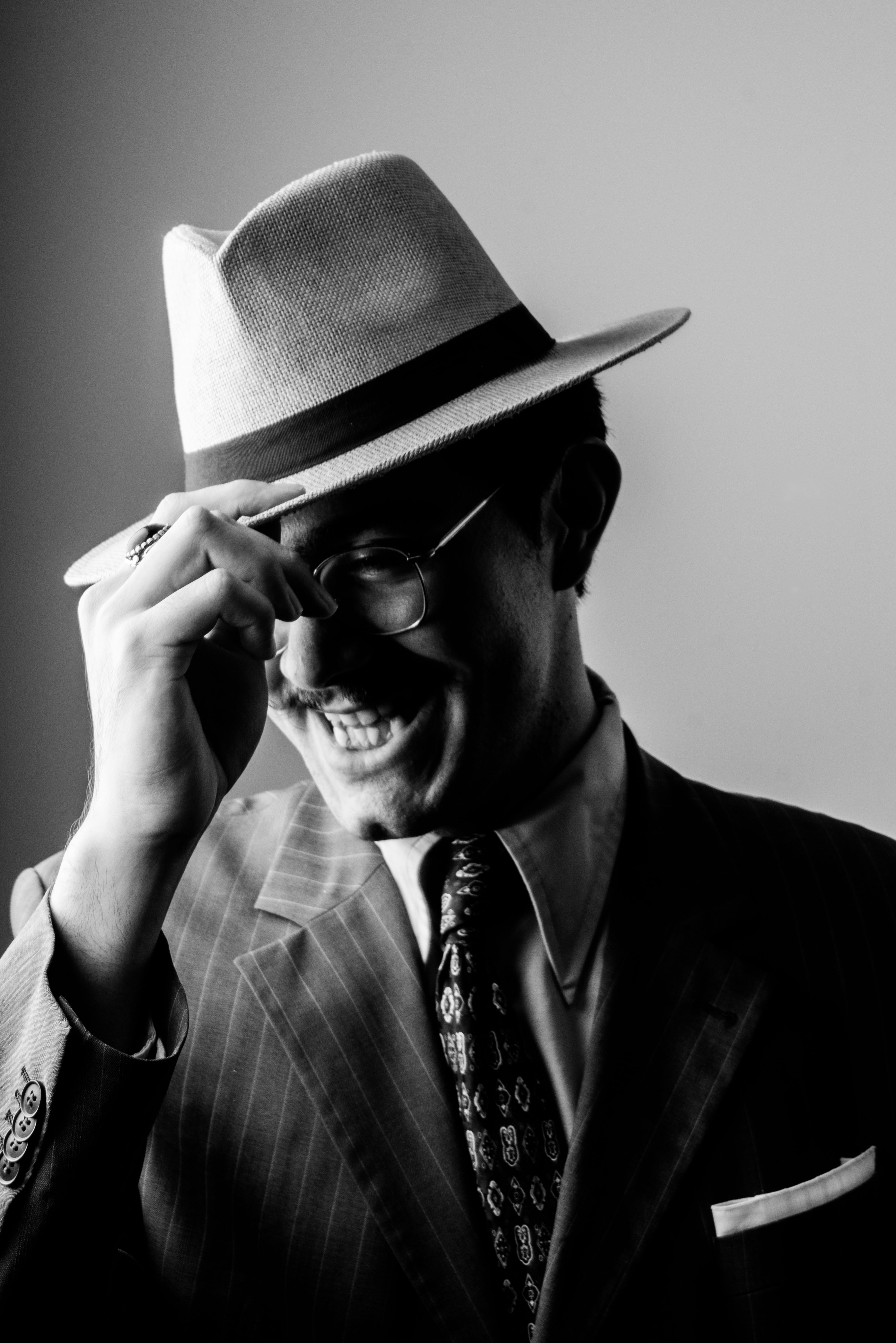 Black and white portrait of a man in vintage clothing tipping a fedora hat with a smile.