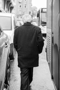 An elderly man in a coat walks between vehicles on a city sidewalk, captured in black and white.